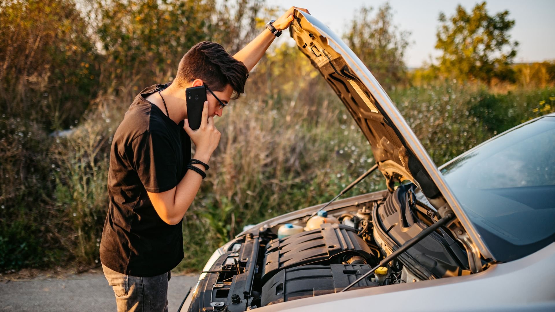 Person on phone next to open car hood, troubleshooting vehicle problem